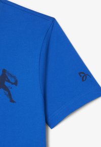 Sininen puuvillapaita, jossa on pyöreä kaulus. Paidassa on musta grafiikka pelaajasta ja musta logo hihassa. Sileä rakenne, lyhyet hihat.