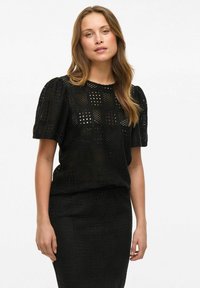 Crop top noir avec manches bouffantes, présentant un design en tricot ouvert texturé et des motifs carrés, assorti d'une jupe noire texturée.