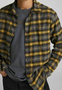 Camicia in flanella a quadri gialli e grigi, abbottonata con tasca sul petto. Indossata sopra una maglietta grigia, presenta maniche lunghe e colletto.