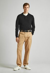 Camisa polo negra de manga larga, pantalones relajados beige con bolsillos frontales y deportivas blancas con acentos negros, de pie contra un fondo neutro.