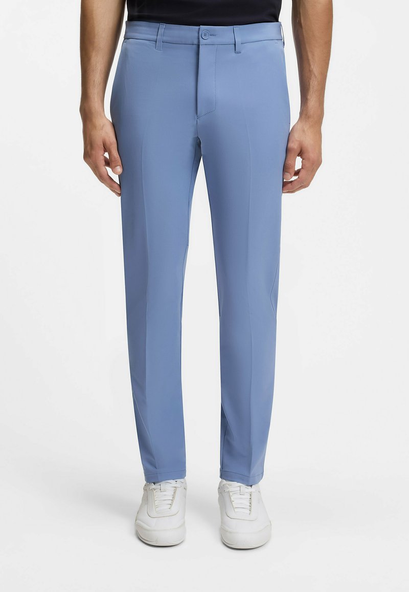 Homme portant un pantalon bleu clair ajusté et des baskets blanches, debout devant un fond blanc, les mains détendues le long du corps.