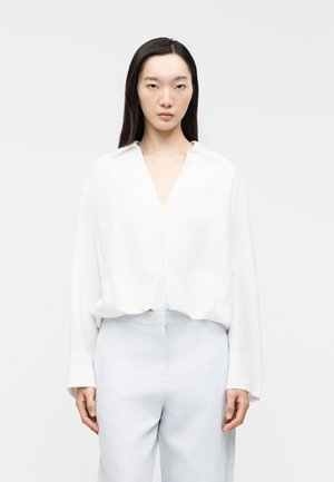 Woman with long dark hair wearing a loose white V-neck blouse and light blue high-waisted pants, standing against a plain background.