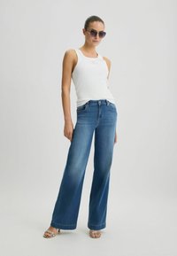 Jean bleu à jambes larges avec un dégradé, associé à un débardeur blanc à côtes. Le modèle porte également des lunettes de soleil noires et des sandales ornées.