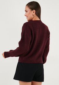 Burgundy színű, textúrált pulóver hosszú ujjakkal és bordázott mandzsettával, fekete rövidnadrággal viselve. Laza szabású és kerek nyakkivágással rendelkezik.