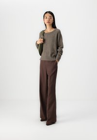 Maglione grigio lavorato a maglia con scollatura ampia, abbinato a pantaloni marroni a vita alta. Il modello tiene una borsa verde. Texture liscia, vestibilità rilassata.