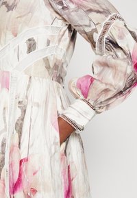 Robe fleurie en tissu léger avec des motifs rose et blanc, dentelle le long des coutures, et manches bouffantes avec des poignets élastiques.