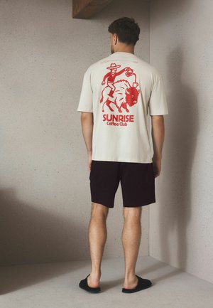 Mężczyzna noszący beżowy t-shirt z czerwonym nadrukiem kowboja przelewającego kawę na bizona i tekstem "SUNRISE Coffee Club,", zestawiony z czarnymi szortami i kapciami.