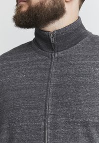 Grå zip-up-jacka med hög krage, tillverkad av en texturerad stickad tyg; har en framficka med dragkedja och en sportig design.