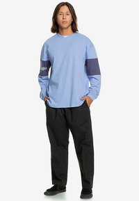 Chemise bleu clair à manches longues avec un accent marine sur les manches, associée à un pantalon noir à motifs. La tenue est complétée par des chaussures noires.