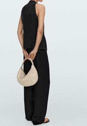 Žena v černém tílku bez rukávů a širokých černých kalhotách drží za zády béžovou pletenou kabelku a stojí na světlém pozadí.