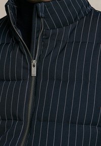 Chaleco acolchado azul marino con rayas blancas verticales, cuello alto y cierre de cremallera plateada. Presenta textura acolchada y secciones acolchonadas.