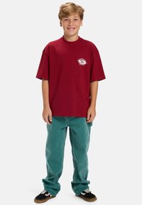 Rode katoenen t-shirt met een kleine witte afbeelding op de borst, gecombineerd met losse teal corduroy broeken en zwarte sneakers.