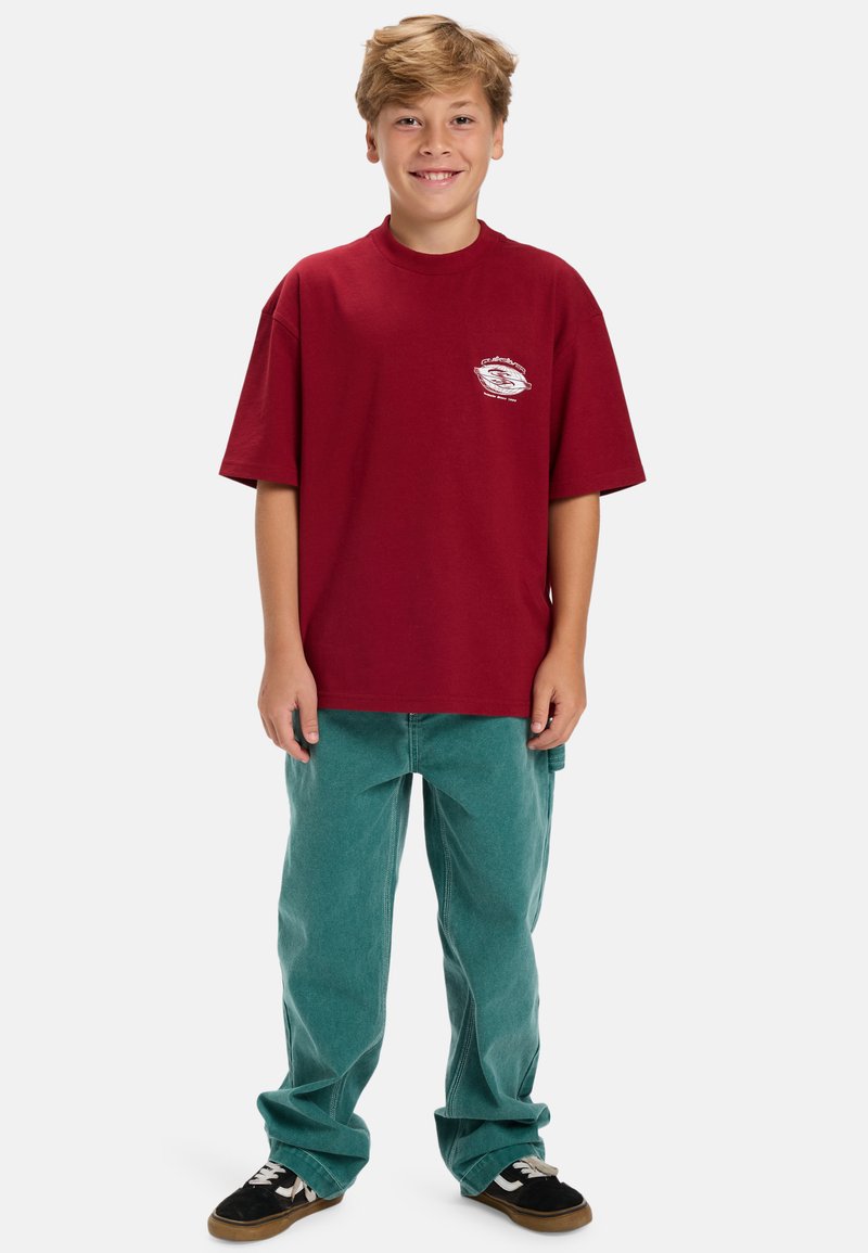 Rode katoenen t-shirt met een kleine witte afbeelding op de borst, gecombineerd met losse teal corduroy broeken en zwarte sneakers.