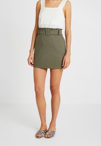 Jupe en coton verte avec une ceinture, design ajusté, assortie à un haut sans manches blanc et des sandales à motifs. Look simple et moderne.