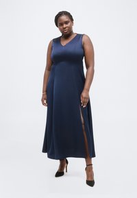 Γυναίκα με αμάνικο μακρύ φόρεμα σε ναυτικό μπλε με πλευρικό άνοιγμα, μαύρα πέδιλα με λουράκι στον αστράγαλο, χρυσό βραχιόλι και μικρά σκουλαρίκια κύκλου, στέκεται με αυτοπεποίθηση.