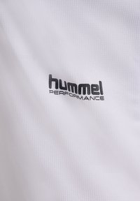 Hvid struktureret stof med sort "hummel PERFORMANCE"-logo trykt på, der viser mærkedetaljen på sportsbeklædning.