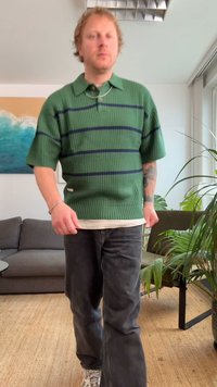 Muž kráčajúci v interiéri, oblečený v zelenom svetri s krátkym rukávom a čiernymi pruhmi, tmavých džínsoch a bielych teniskách, v prostredí obývačky.