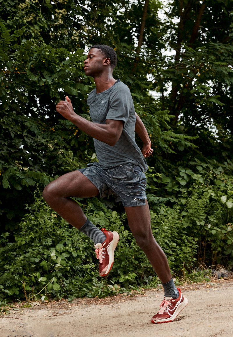 Ein Athlet in einem grauen, strukturierten Shirt, gemusterten Shorts und roten Laufschuhen mit weißen Akzenten, der auf einem Weg in der Nähe von Grünflächen sprintet.