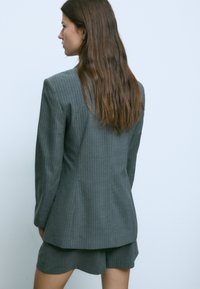 Sivi blazer z modrimi črtami z oblikovano zasnovo in dolgimi rokavi, združen z ujemajočimi se kratkimi hlačami, pogled od zadaj. Gladek tekstura.