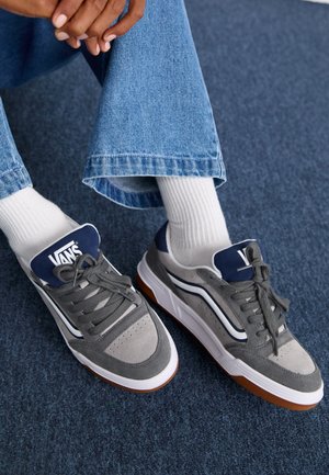 Graue Sneaker mit marineblauen Akzenten, Wildleder- und Texturpanels, weißen Schnürsenkeln und Gummisohlen. Weiße Kniesocken und Jeans sind sichtbar.