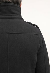 Personne portant un manteau en laine noir avec un col haut et une épaulette, vue de dos sur un fond uni.