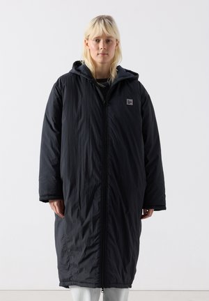 Nainen, joka pukeutuu pitkään mustaan hupulliseen vetoketjulliseen takkiin, seisoo valkoisen taustan edessä kasvot kohti kameraa.