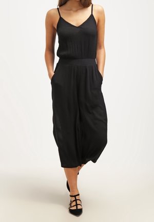 Kobieta w czarnym, bezrękawnym kombinezonie z szerokimi nogawkami i kieszeniami, ubrana w czarne sandały na paski, stojąca na tle jasnego, jednolitego tła.