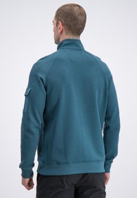 Turkusowa bluza na zamek z teksturowanej tkaniny, z wysokim kołnierzem i ściągaczami na rękawach. Posiada boczną kieszeń na lewym rękawie.