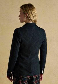 Námornícky herringbone blejzer s prispôsobeným strihom, jednoradovým zapínaním, špicatými klopami a perlami na gombíkových akcentoch. Viditeľné nohavice s kontrolným vzorom.