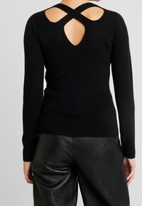 Svart långärmad tröja gjord av mjukt material med ett korsningsdesign bak, med en nyckelhålsdetalj. Accenterad med en slät textur.