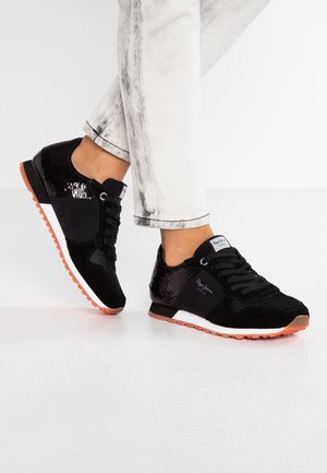 Zapatillas negras con lentejuelas y suelas naranjas, usadas con vaqueros lavados claros sobre un fondo de suelo blanco.