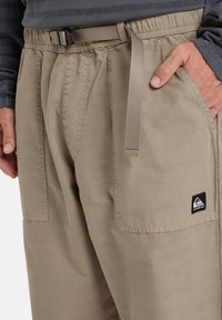Beige cargo pants with a drawstring waistband. Features front pockets and a black logo patch on the lower leg. Soft, woven fabric texture.