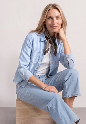 Femme aux cheveux blonds assise sur une caisse en bois, vêtue d'une veste et d'un pantalon en denim bleu clair, d'une chemise blanche et d'un foulard à motifs autour du cou.