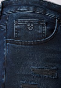 Granatowe dżinsy mają wyraźne przetarcia, małą kieszonkę na monety i postrzępione dziury w pobliżu głównej tylnej kieszeni. Logo wytłoczone na materiale.