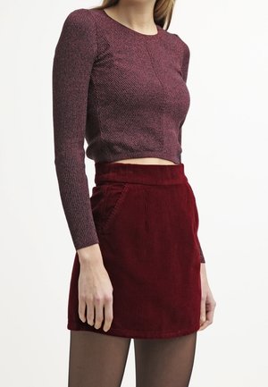Kvinde iført en langærmet, tekstureret bordeaux crop top og en rød fløjlsmini nederdel med lommer, stående mod en enkel hvid baggrund.