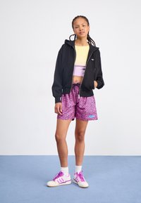 Felpa nera con zip, top corto pastello giallo e viola, pantaloni corti leopardati rosa, scarpe bianche con dettagli rosa, posizionati su un pavimento blu.