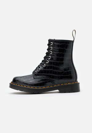 Bottine noire brillante en cuir à motif crocodile, lacets, coutures jaunes et languette arrière sur fond uni.