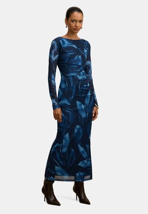 Naine seisab, kandes pika varrukaga, keha järgiva lõikega tumel sinist kleiti, millel on suured sinised lehemustrid, ning pruunide kontsadega pahkluu saabastega.