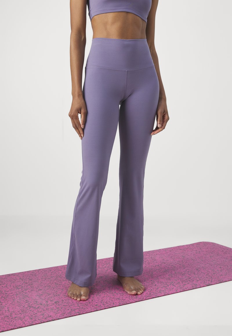 Pantalones de yoga morados acampanados hechos de tela elástica, con una cintura alta. Esterilla de yoga texturizada rosa debajo. Pies descalzos visibles.