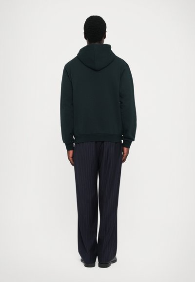 Dark green hoodie with a lined texture, featuring a large hood and ribbed cuffs, paired with loose-fitting navy pinstriped trousers.
