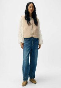 Beige fleecevest met ronde hals en drukknoopjes over een wit gestructureerd shirt met lange mouwen, gecombineerd met losse blauwe jeans en beige schoenen.
