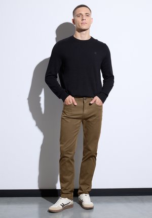 Hombre de pie contra una pared blanca, vistiendo un suéter negro de manga larga, pantalones marrones y zapatillas blancas, con las manos en los bolsillos.