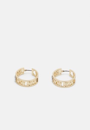 Pari kultaisen sävyisiä renkaanmuotoisia korvakoruja, joissa on leikattu DKNY-logo ja metalliset nastat valkoisella taustalla.