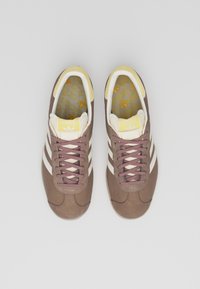 Zapatillas deportivas de ante marrón con franjas crema y detalles amarillos. Punta redonda, diseño con cordones y plantillas estampadas con el logo de Adidas.