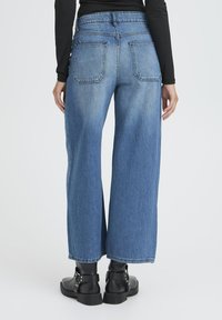 �Široke modre jeans hlače z visokim pasom, opremljene z dvema zadnjima žepoma in raztrganim robom. Kombinirane z črnimi gležnjači.