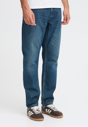 Une personne porte un jean bleu à jambes droites, une chemise blanche et des baskets marron sur un fond blanc uni.