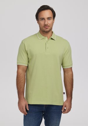 Mężczyzna z krótkimi ciemnymi włosami, ubrany w jasnozieloną koszulkę polo z krótkim rękawem i niebieskie dżinsy, stojący na tle jednolitego jasnego tła.