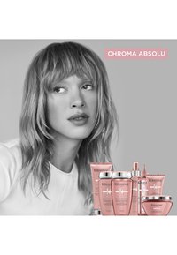 Kvinde med mellemlangt bølget hår ser til siden; pink Kérastase Chroma Absolu hårplejeprodukter vist nedenfor hende på grå baggrund.