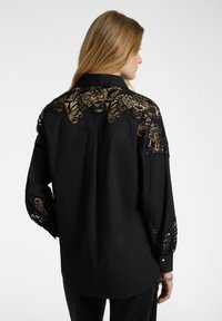 La blusa nera presenta dettagli in pizzo nero intricati sulle spalle e sulle maniche, con colletto e polsini abbottonati. Texture setosa.