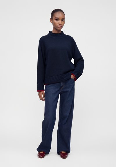 Femme debout, une main dans la poche, portant un pull bleu foncé, un jean à jambes larges et des chaussures rouges sur un fond gris uni.